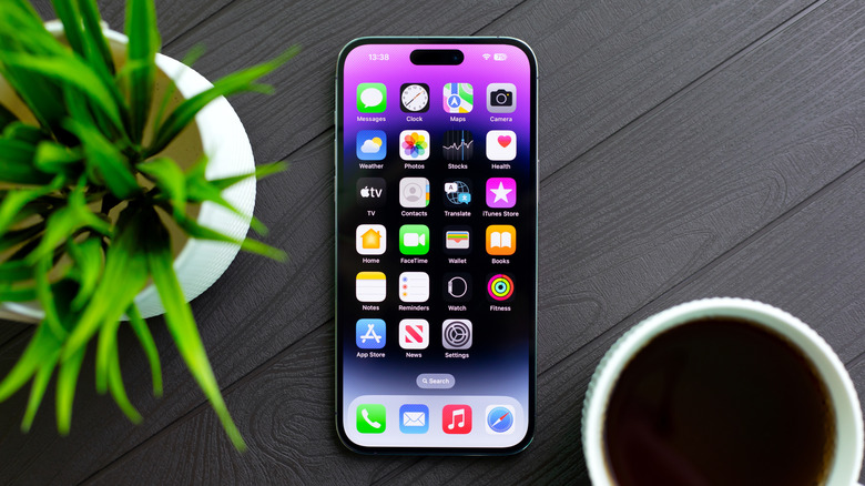 An iPhone displaying its home screen sitting on a wooden table in between a plant and a cup of coffee
