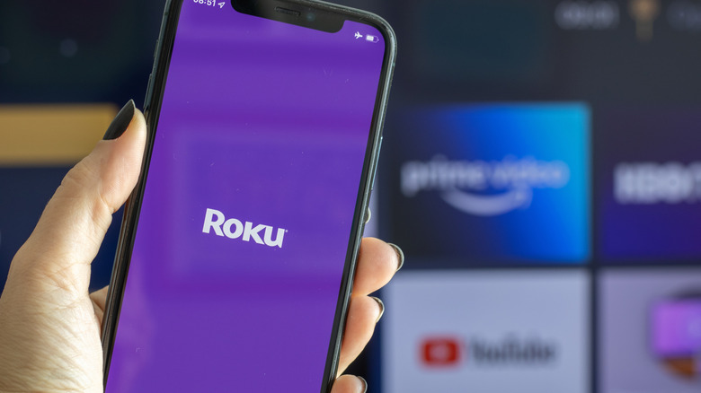A smartphone displaying the Roku logo on its screen with a TV turned on showing Roku UI in the background