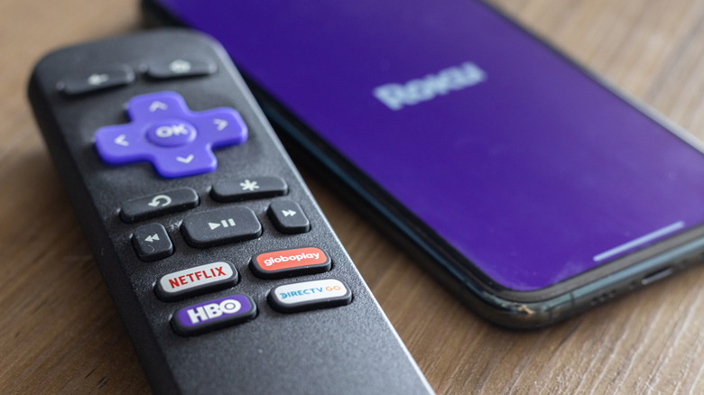 A Roku remote lying beside a smartphone that's displaying the Roku icon