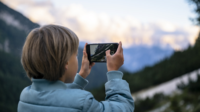 Over the shoulder view of a boy taking a picture with his smart phone of beautiful mountains while he is taking a break during hiking.