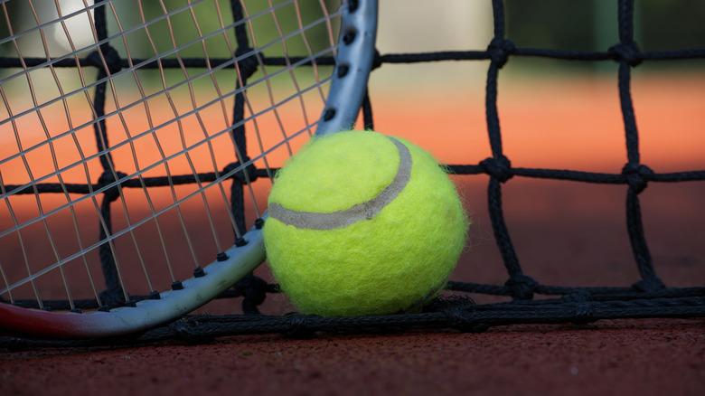 A tennis ball and tennis racket leans against a tennis net on a court