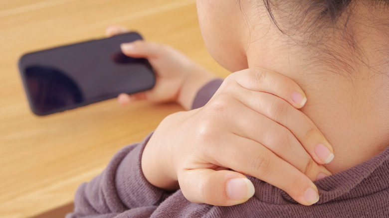 a woman rubbing at her neck pain while looking at her smartphone