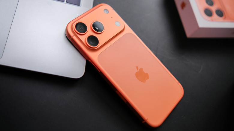 A Cosmic Orange iPhone 17 Pro partially sits on the corner of a silver MacBook, the box for the iPhone can barely be seen in the upper-right corner of the image