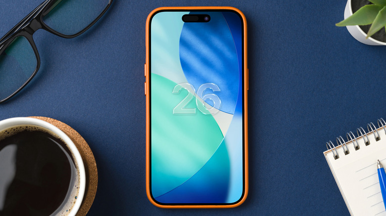 An iPhone with an orange case displays a mockup of iOS 26 on its screen, the iPhone is surrounded by a coffee cup, a small plant, a notepad, and a pair of glasses