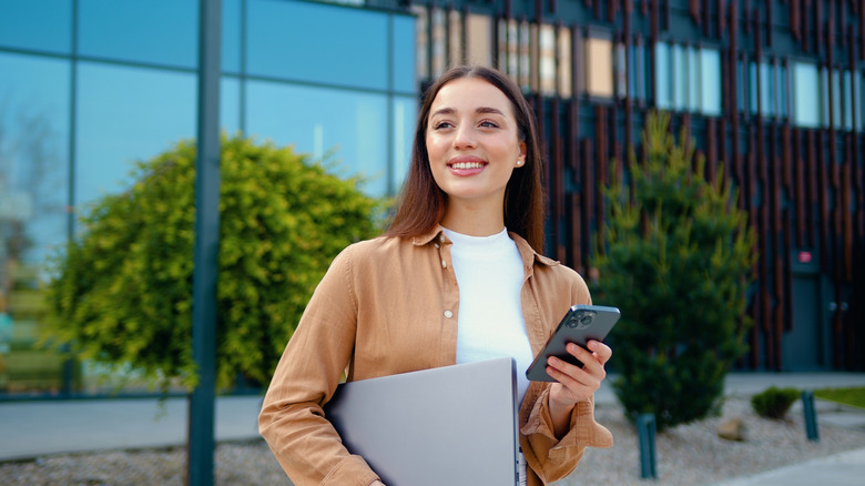 A woman outside holding a phone and a laptop and smiling.