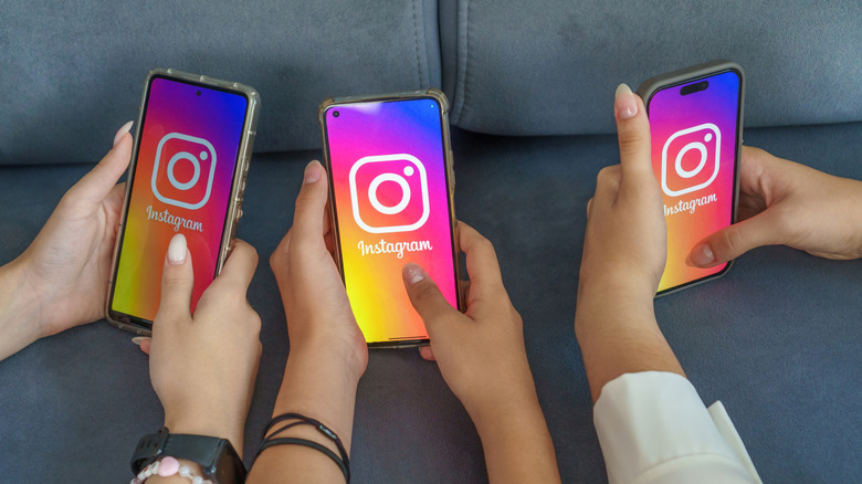 Three individuals using a couch to rest their hands while they hold smartphones displaying the Instagram logo in their hands, the hands are the focal points of the photo and you can not see anyone's face or body