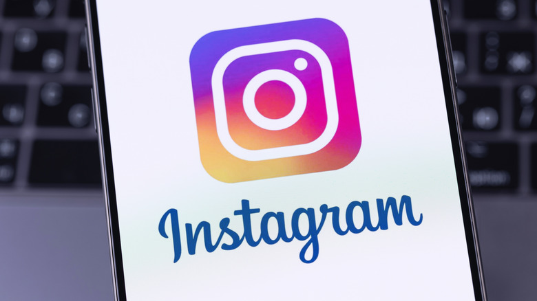 The Instagram logo appears on a smartphone screen, a blurry MacBook keyboard is behind it