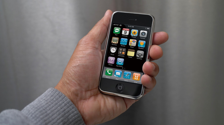 A person's hand holding a 1st generation iPhone with the main display turned on.