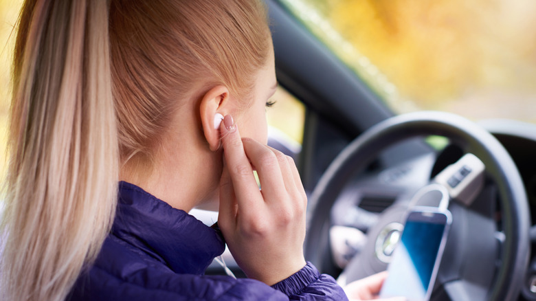 Woman sitting in driver's seat of car adjusting AirPod in ear and holding phone