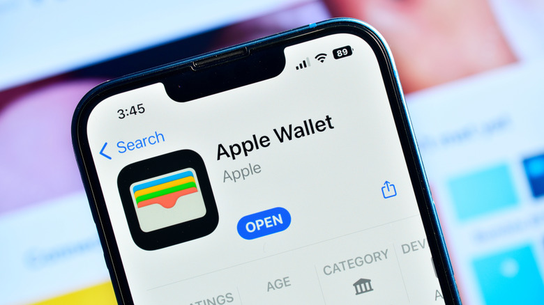 An iPhone displaying the Apple Wallet app within the Apple App Store, behind the device is a screen that is displaying information far too blurry to be read clearly