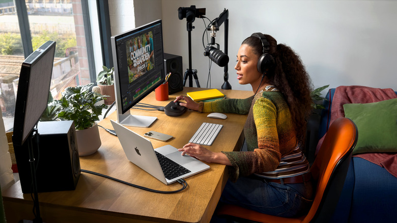 A young woman wearing headphones speaks into a podcast mic while working on a MacBook Pro hooked up to a computer monitor at a desk