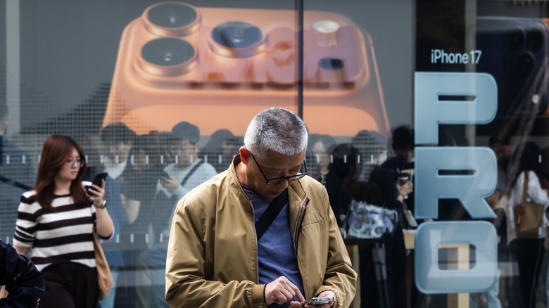 A person using a smartphone in front of an Apple retail store featuring iPhone 17 Pro signage.