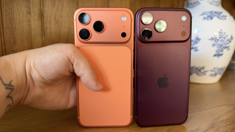 iPhone 18 Pro Max and 17 Pro Max stood next to each other on wood sideboard with hand grabbing the 17 Pro Max