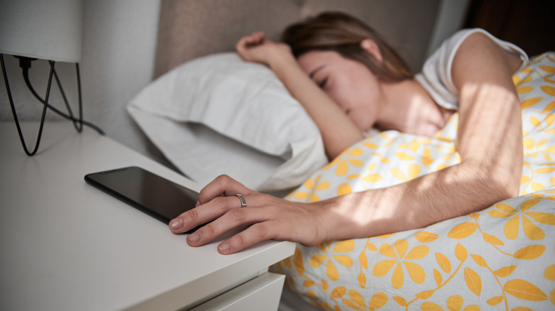 A young woman in bed reaching for a smartphone.