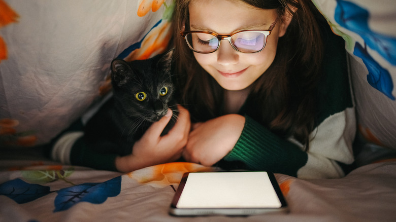 Child reading on a Kindle under a blanket while holding a black cat.
