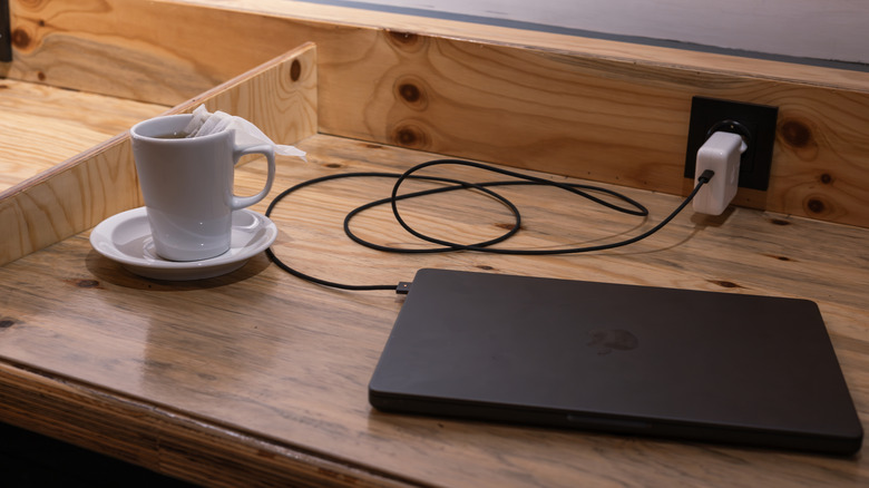 A MacBook charger is connected to a wall outlet, which is connected to a black, closed MacBook. The laptop is on a wooden table, next to a mug of tea.