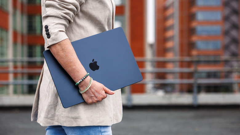 A person carrying a closed-up MacBook with a blurred city setting in the background.