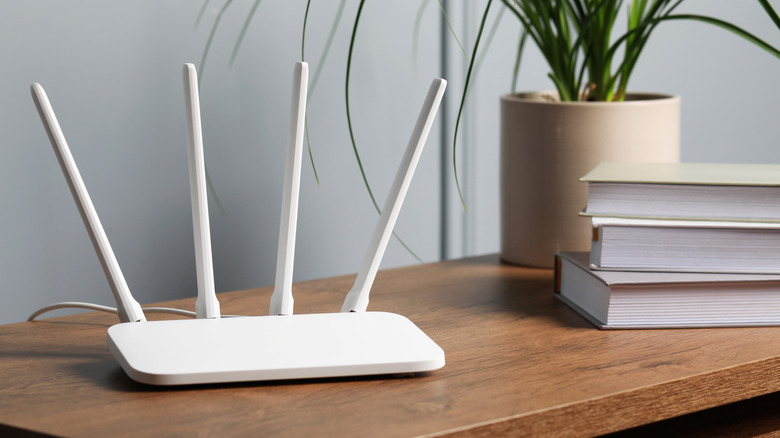 A white router with four antennas on a wooden table alongside a stack of books and a plant