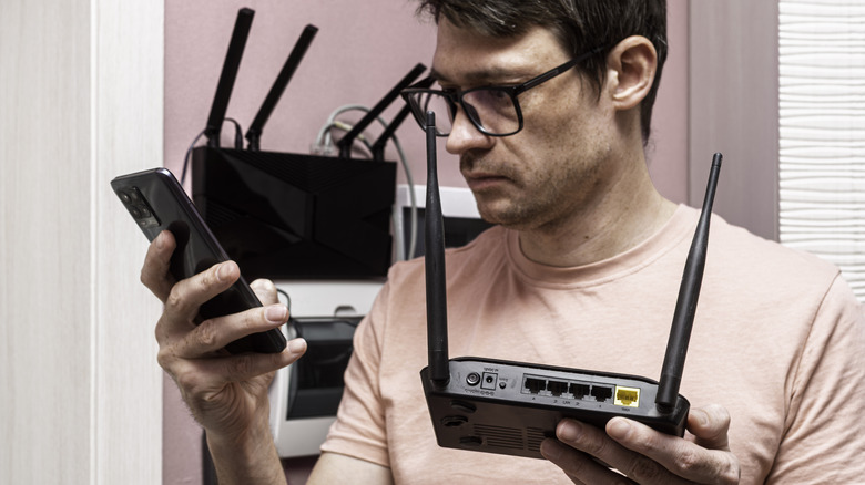 User holding a phone in one hand and wireless router in the other with more network equipment in the background.