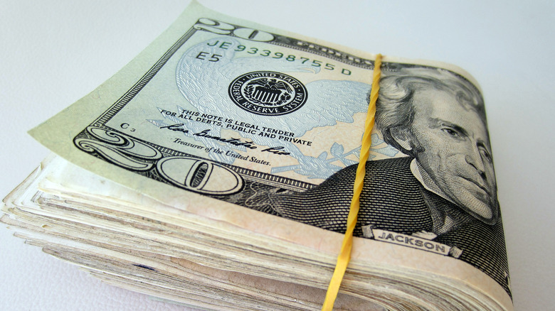 A wad of American cash bills wrapped in a yellow rubber band, with one 20-dollar bill on the outside.