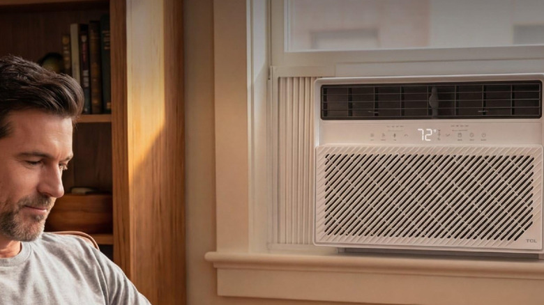 A man sitting in a living room, next to a window-mounted TCL air conditioner.