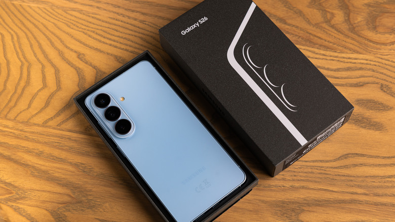 A blue Samsung Galaxy S26 still sitting in its box with the top portion of the box sitting next to the device, both items are sitting on a wooden surface