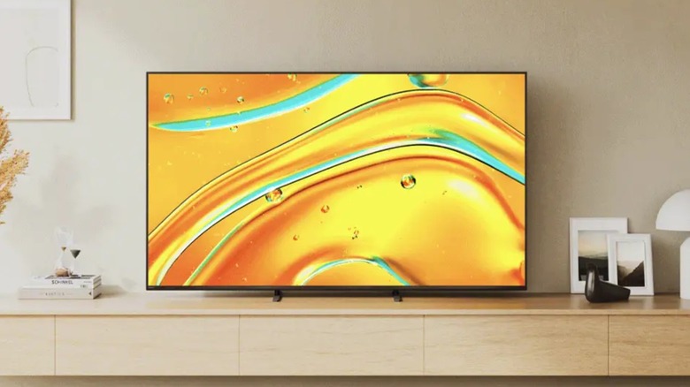 A Sony Bravia 5 Google TV displaying a kaleidoscope of yellow and blue colors sits on a TV shelf amongst some photo frames, a lamp, and two small books