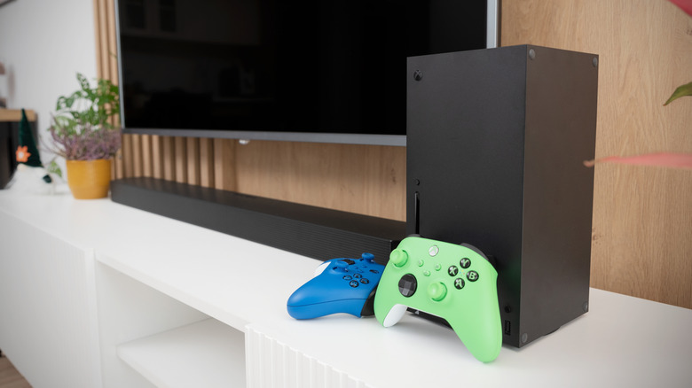 An Xbox Series X with two controllers on a stand beside a TV