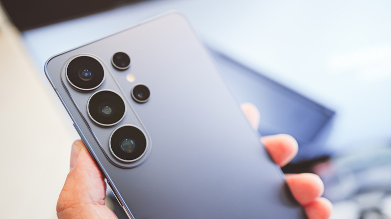 The backside of a silver Galaxy S26 Ultra as it sits in a user's left hand, the background is far too blurry to establish the location of the image