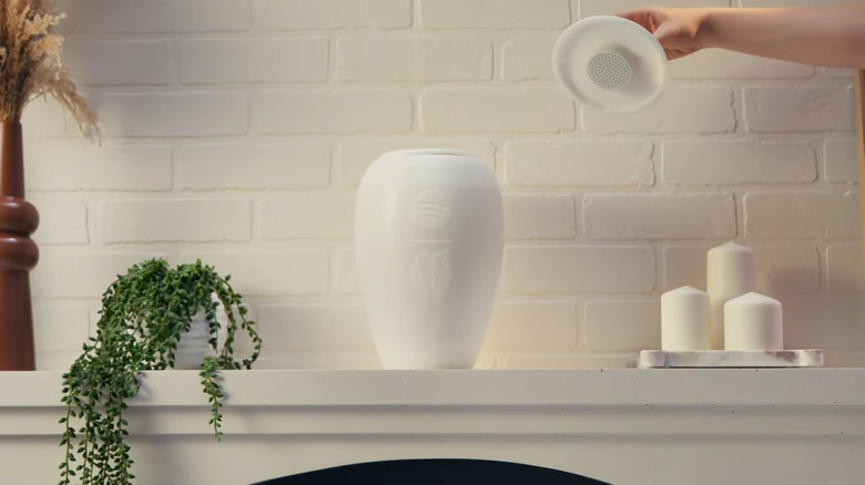A customer lifts the top off of the Eternal Playlist Urn as it sits prominently on a white mantel beside flowers an candles.