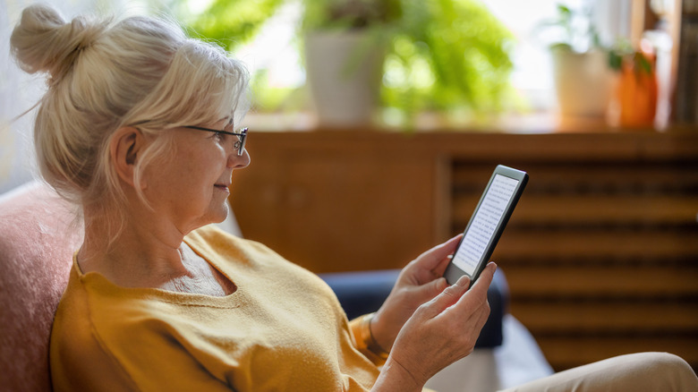 An older woman with glasses looks smilingly on her Kindle as she reads it on her couch