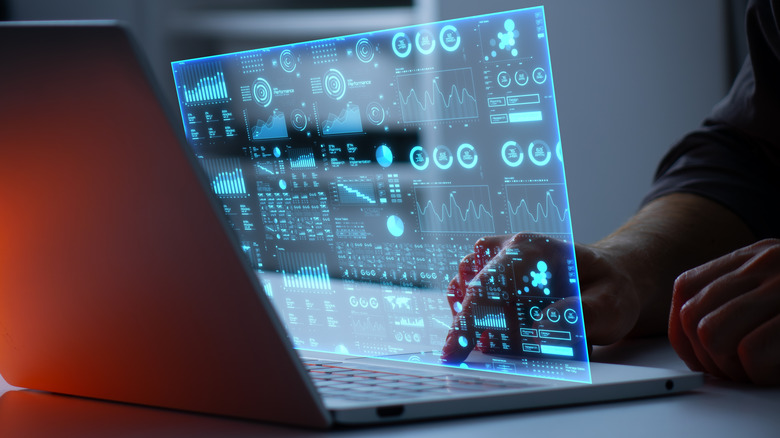 A laptop sits on a desk, a holographic display full of data and graphs floats over the keyboard.