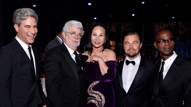 Leonardo DiCaprio and George Lucas stand with a group of smiling acquaintances at a black tie event.