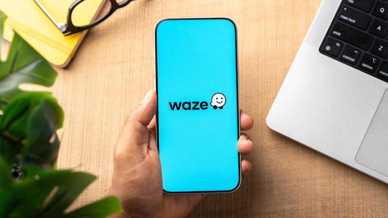 Hand holding a smartphone displaying the Waze app logo on a blue screen, placed on a desk with partially visible laptop, notebook, glasses, and plant.