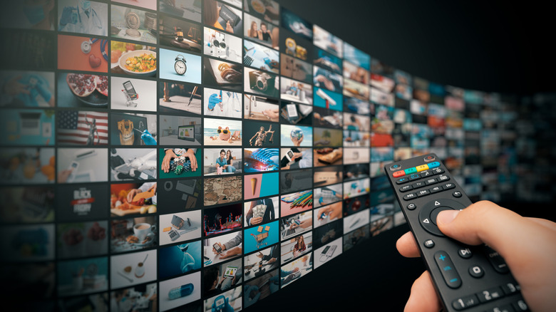 A hand using a remote controller to select a TV show or movie from a huge display wall of available content.
