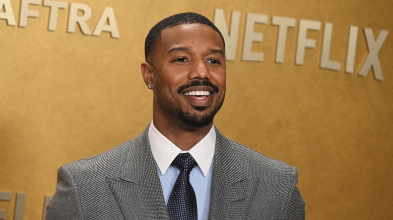Actor Micheal B. Jordan in a grey suit with a blue tie stands smiling in front of a gold backdrop displaying the NetFlix logo