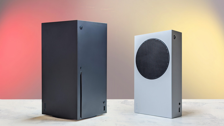 A black Xbox Series X stands next to a white Xbox Series S, both consoles are standing on what appears to be a white marble surface in front of a colorful backdrop