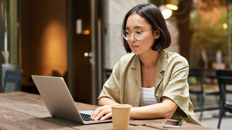A woman working on a laptop at an outdoor café table, with a coffee cup and smartphone nearby