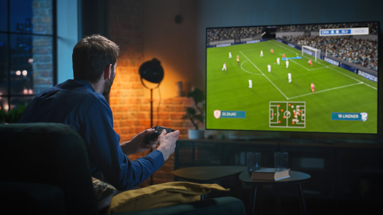 Person playing a soccer video game on a large TV in a dimly lit living room.