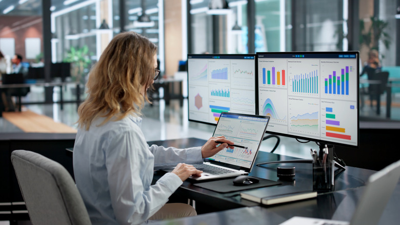 Office worker reviewing data dashboards on a laptop and dual monitors in a modern workspace.