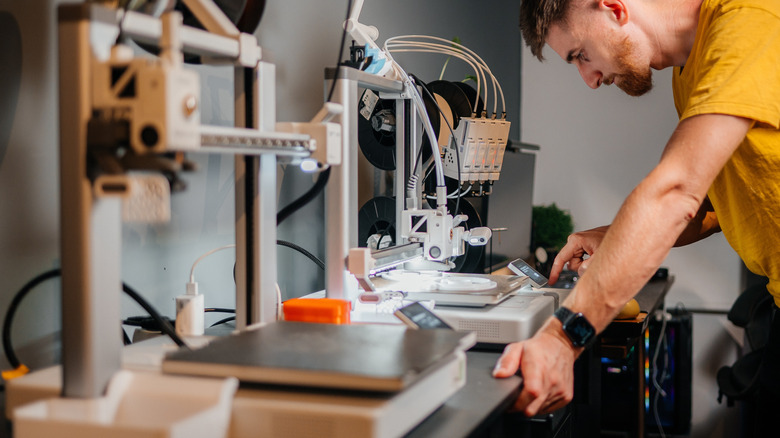 Person operating and adjusting a desktop 3D printer, with filament spools and printing components visible on a workbench.
