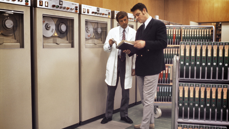 Two men standing beside large mainframe computer tape drives, reviewing notes or documentation in a data center environment.