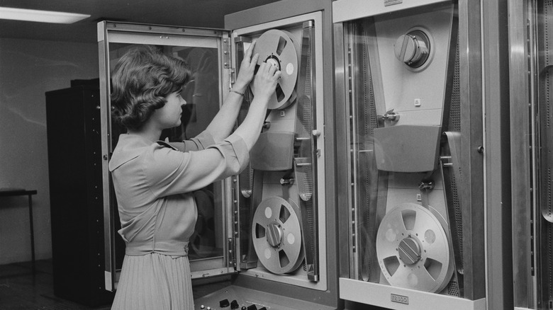 Black-and-white photo of a person loading a large reel of magnetic tape into an early mainframe computer, with tall cabinet units and multiple tape drives visible.