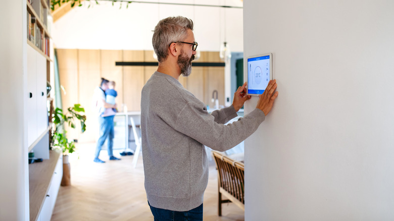 Man adjusting a wall-mounted smart thermostat touch screen in a modern home, with a family scene visible in the background.