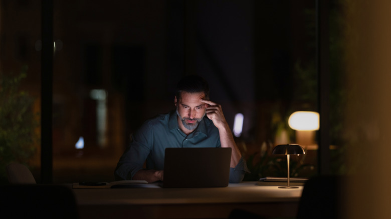 Someone working on a laptop at a desk in the dark with a dim lamp nearby.