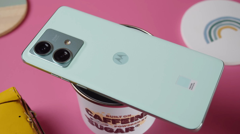 A light green Motorola smartphone sits on top of a coffee cup, both of which sit on a pink backdrop
