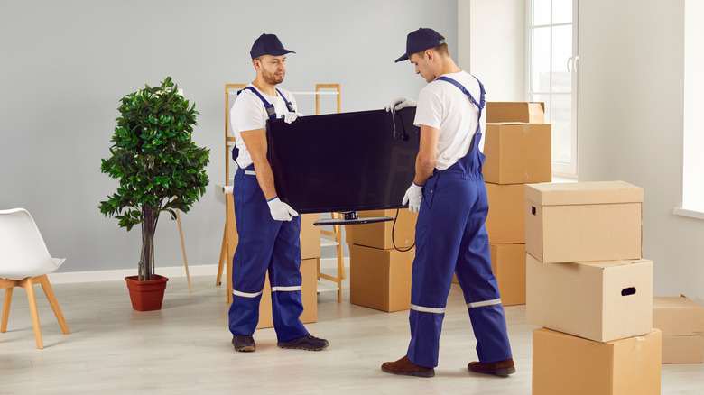 Two movers carrying a flat-screen TV in a room with moving boxes.