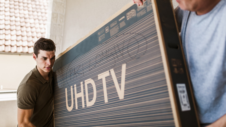 Two people carrying a large UHD TV box indoors.
