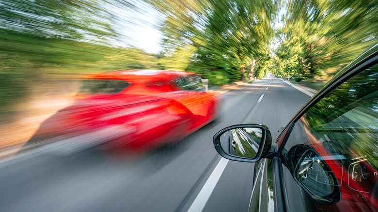 A red car speeds past a black vehicle's side mirror on a tree-lined road.