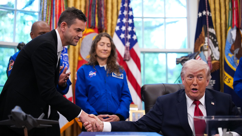 A seated President Trump shakes hands with Jared Isaacman as Artemis astronauts stand at attention in the background